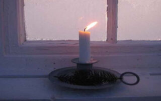 A lit white candle placed on a metal holder near a frosty window, symbolizing hope, inner light, and resilience during uncertain times.