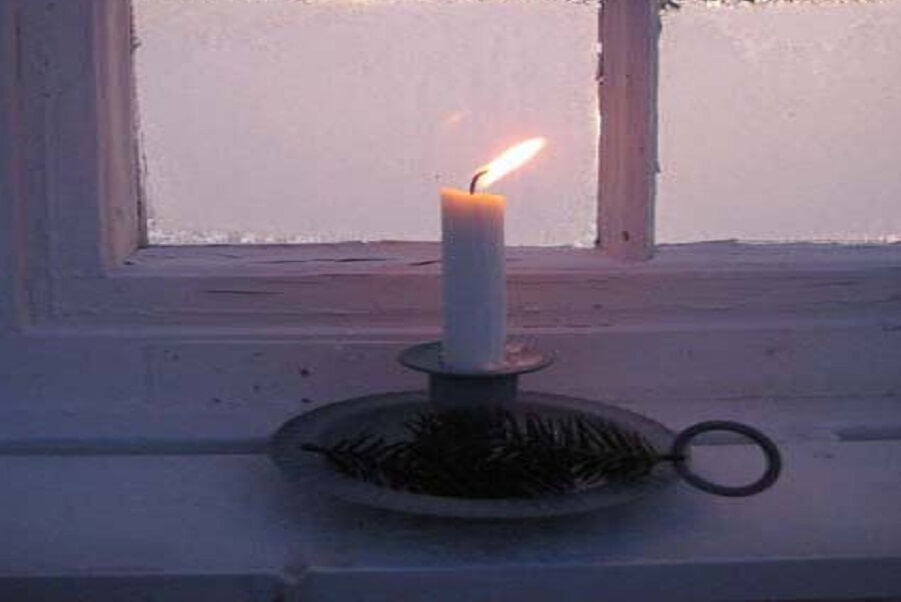 A lit white candle placed on a metal holder near a frosty window, symbolizing hope, inner light, and resilience during uncertain times.