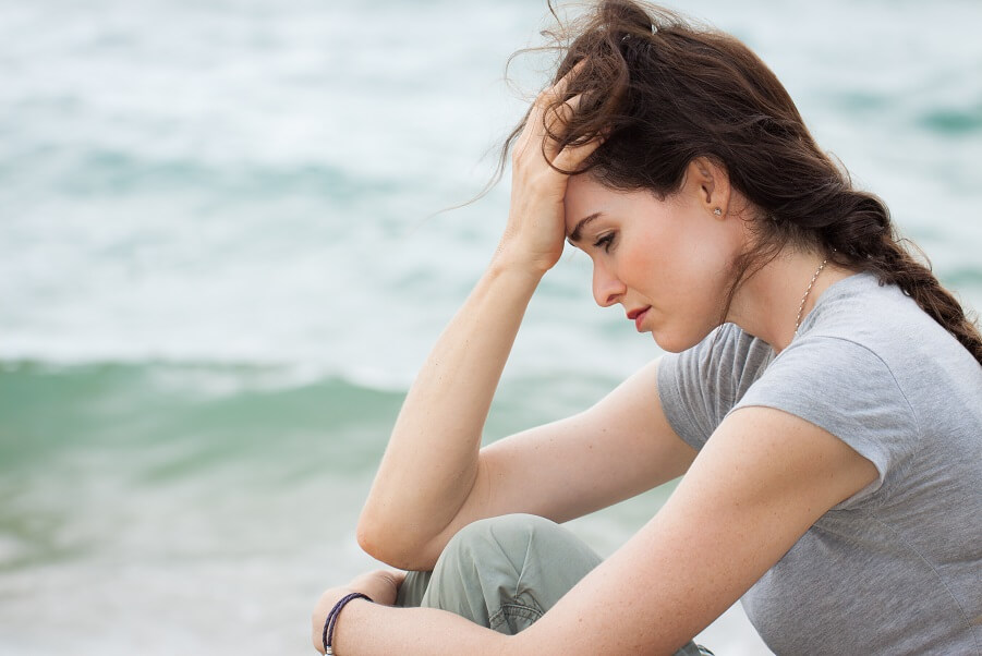 Woman sitting by the ocean looking worried, symbolizing how fear and anxiety pull us away from the present moment