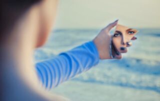 Woman holding a mirror reflecting her face while standing near the ocean, symbolizing self-worth, self-reflection, and personal value.