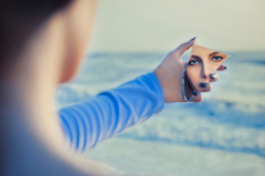 Woman holding a mirror reflecting her face while standing near the ocean, symbolizing self-worth, self-reflection, and personal value.