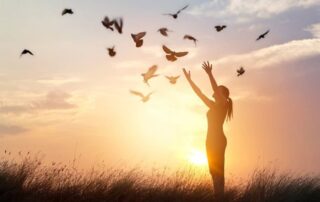 Silhouette of a woman releasing birds into the sunset sky symbolizing letting go, freedom, and emotional healing