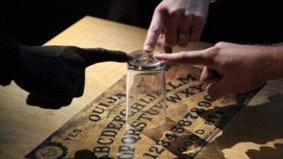 Ouija board session showing human hands and a dark entity hand guiding the glass.