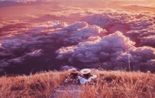 Two women in sun hats sit on a mountaintop trail overlooking pink and purple clouds at sunset.