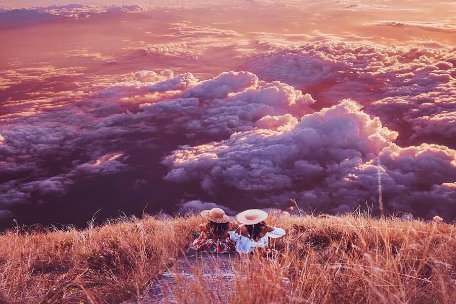 Two women in sun hats sit on a mountaintop trail overlooking pink and purple clouds at sunset.